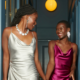 Two women in cocktail dresses laugh and hold hands as they walk down a luxury hallway.