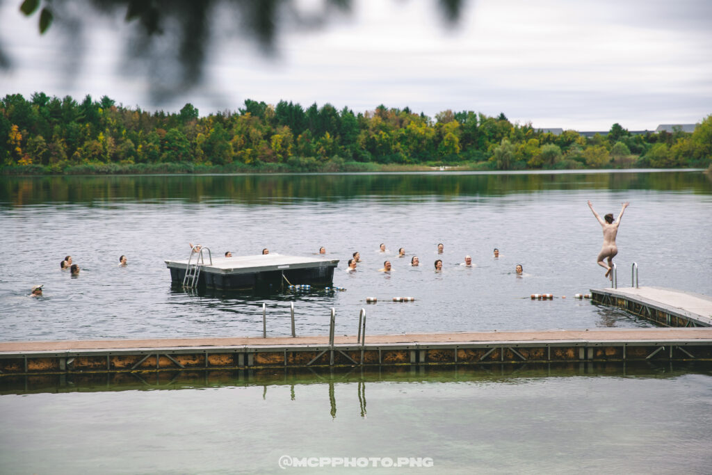 A bunch of naked campers skinny dip in the lake on a gray-sky afternoon.