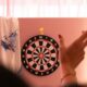 A woman's finger points off to the side as a dart lands in a dart board across the room.