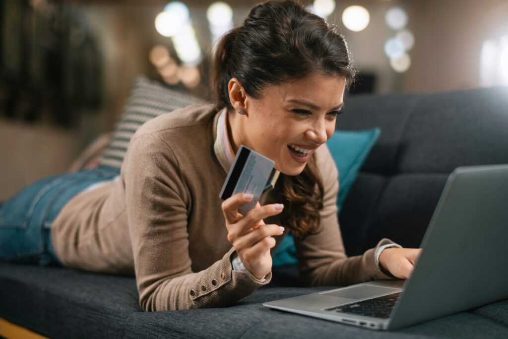 A woman lays on her couch smiling as she holds a credit card in one hand and is scrolling her laptop with the other.