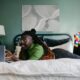 A woman lies on her stomach on her bed, smiling as she looks at her laptop screen.