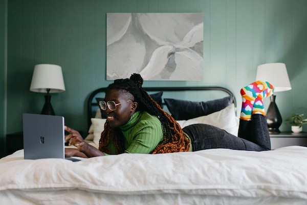 A woman lies on her stomach on her bed, smiling as she looks at her laptop screen.