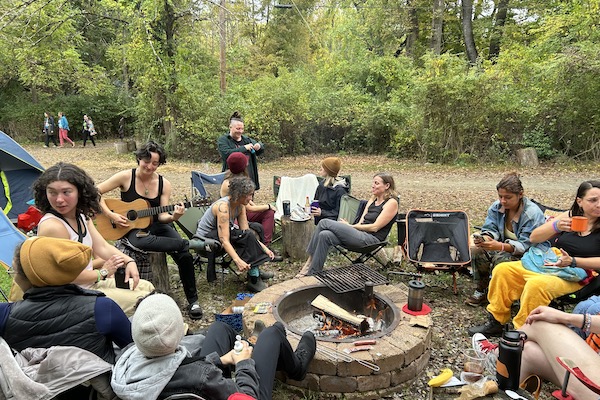 A group of women sit around a campfire in the woods.