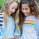 Two young girls huddle together to look at a cell phone.