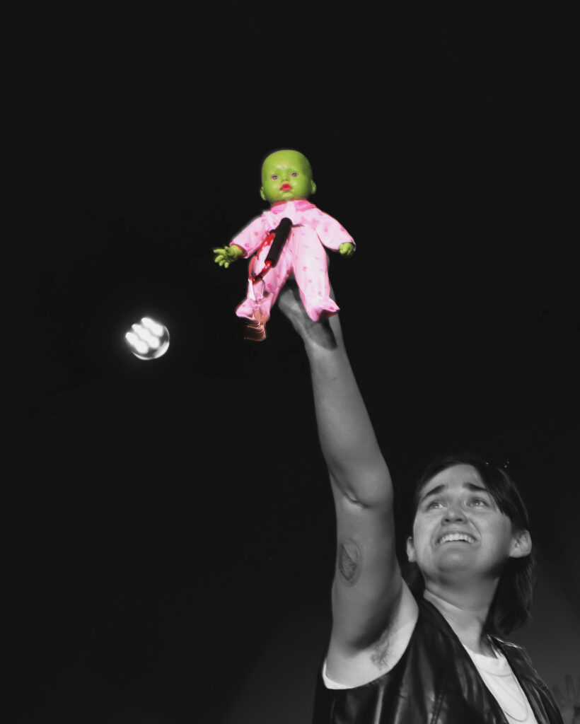 A butch performer holds up the baby Elphaba doll.