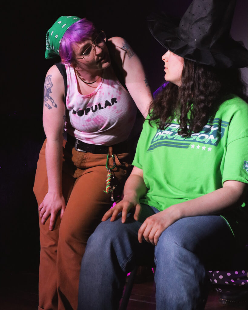 A butch performer in a pink tank top that reads "Popular" interacts with a butch performer in a green top.