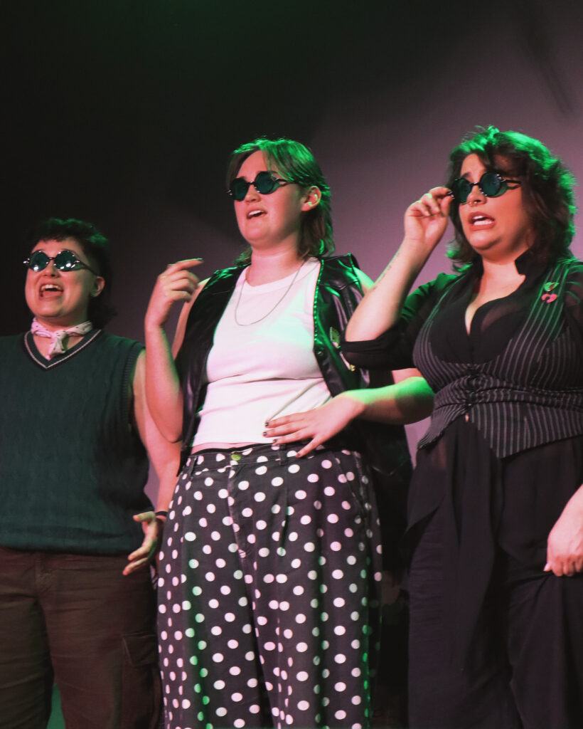 Three butch performers in black and white outfits wear sunglasses as they perform on stage.