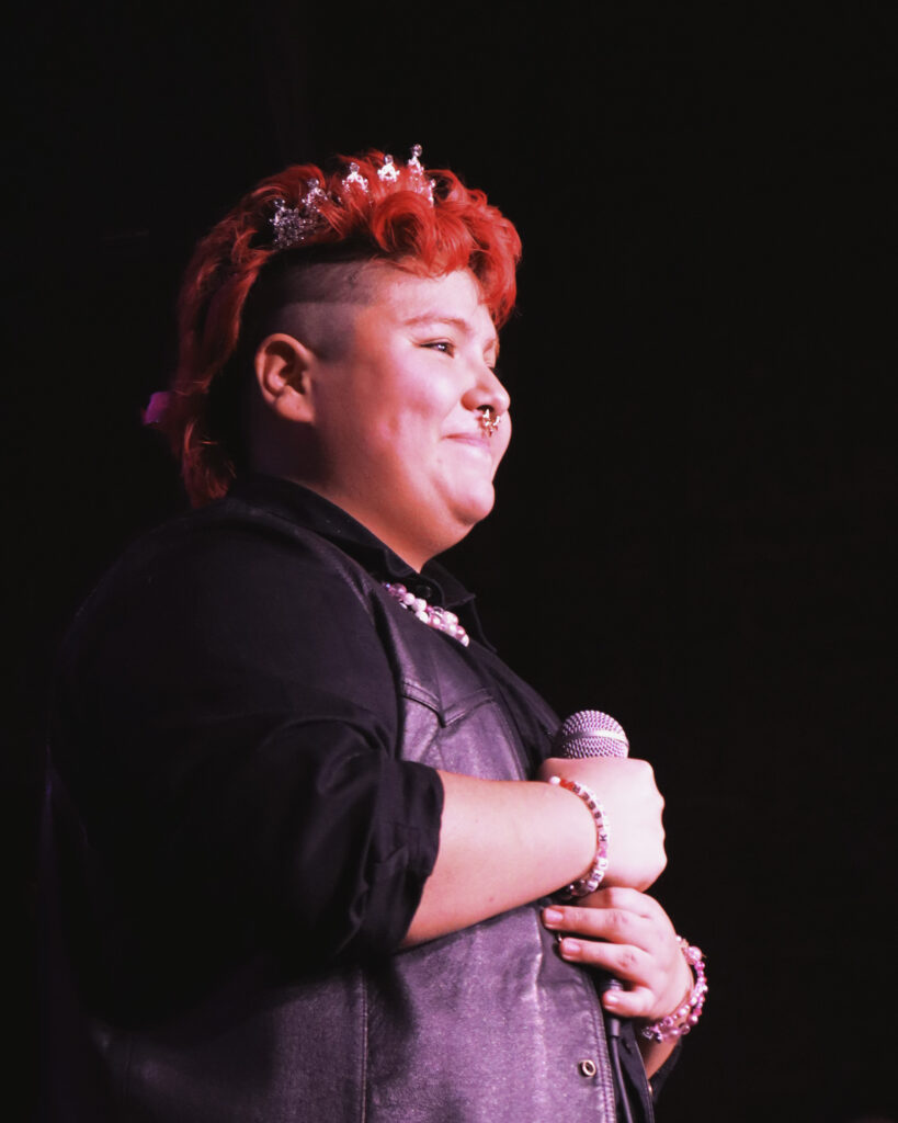 A butch performer in a tiara smiles on stage.