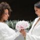 Two women in white—one in a dress and one in a suit—face each other while holding a bouquet.