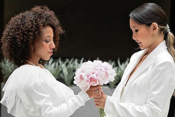 Two women in white—one in a dress and one in a suit—face each other while holding a bouquet.