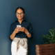 A woman leans against a wall and smiles as she reads something on her phone.