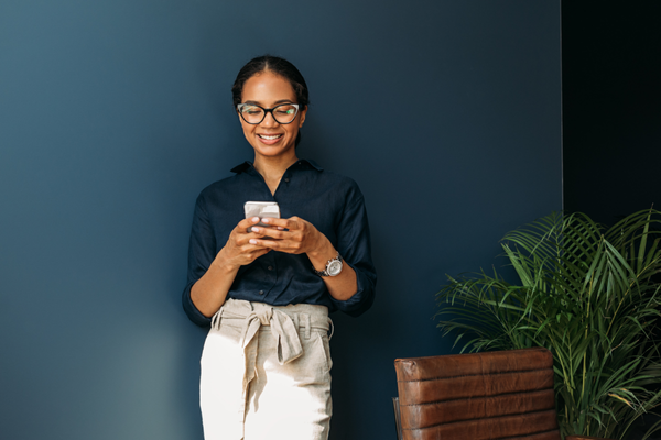 A woman leans against a wall and smiles as she reads something on her phone.