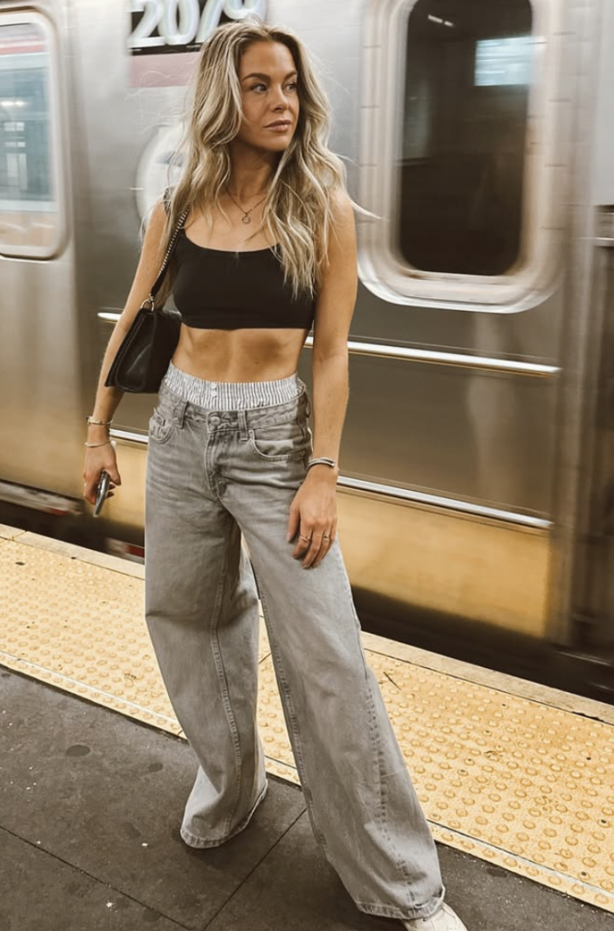 Stacy Synder poses on a subway platform in New York City. She has long blonde hair, wears a black sports bra, and bootleg jeans with boxers peeking out of the top.