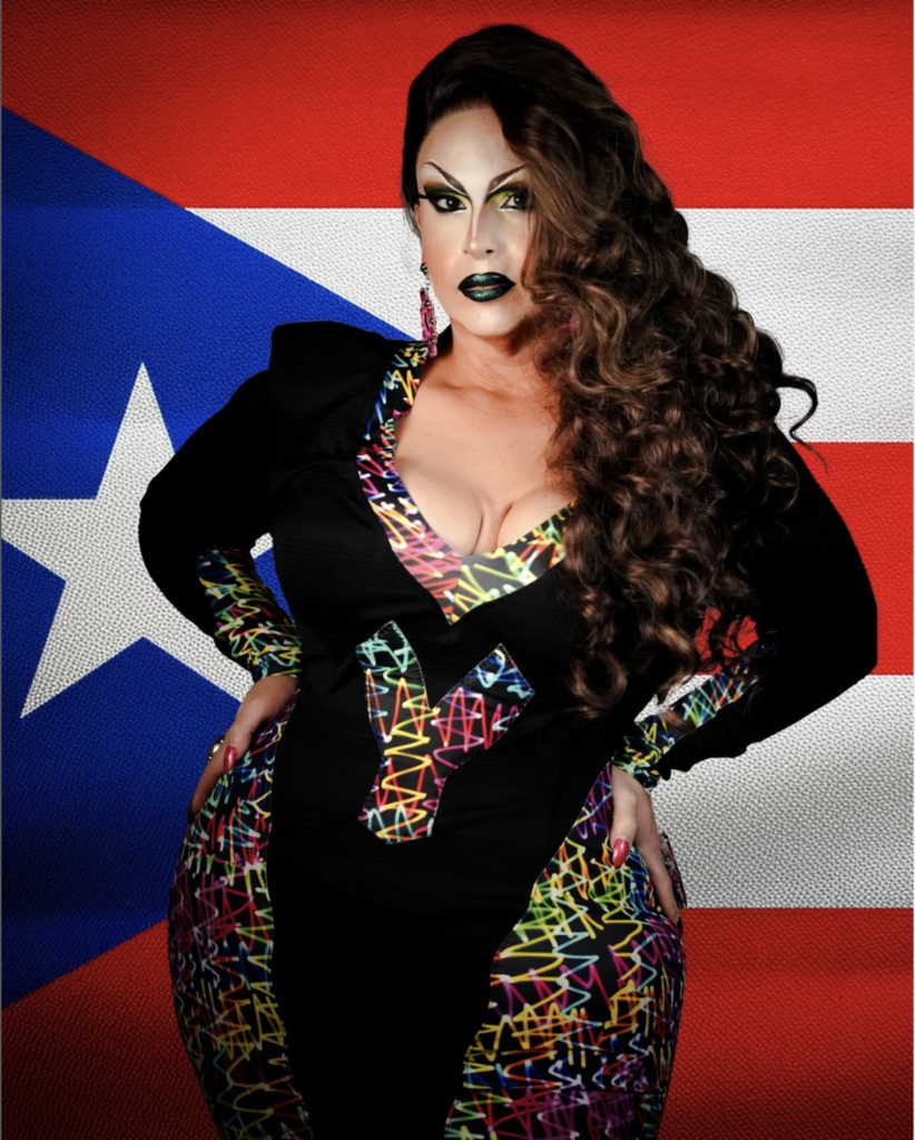 Madame La Queer poses in a black and neon dress in front of the Puerto Rican flag.