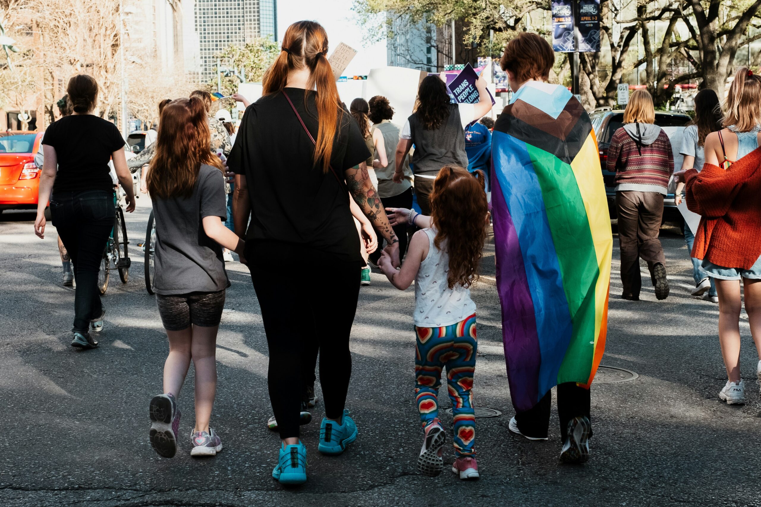 A family of four walks down the street at an LGBTQ event. We see them from behind, walking away. One parent has a pride flag around their shoulders like a cape. One child has rainbow pants on.