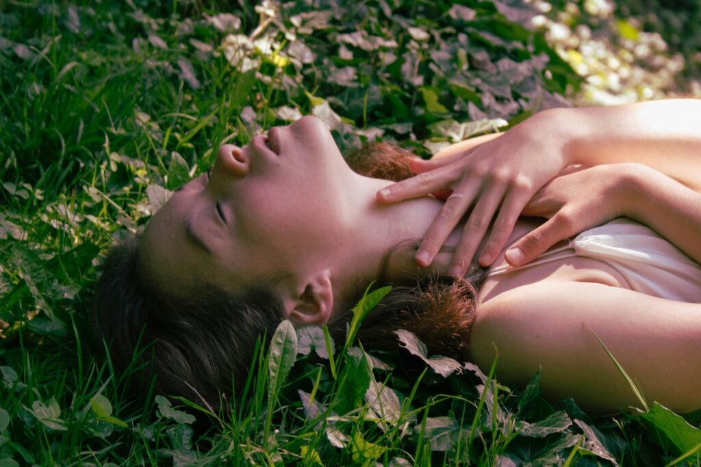 A woman lies on her back in grass, her hands are up around her neck and her eyes are closed.
