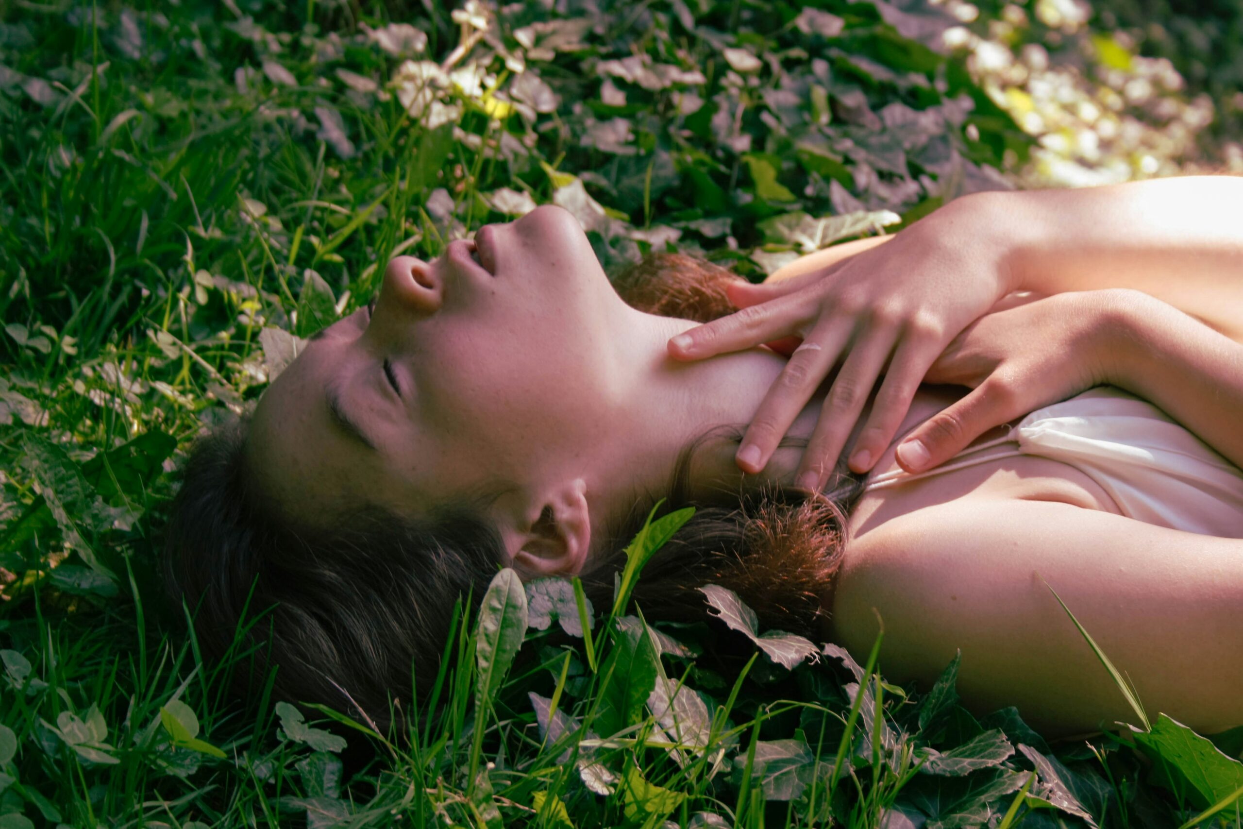 A woman lies on her back in grass, her hands are up around her neck and her eyes are closed.