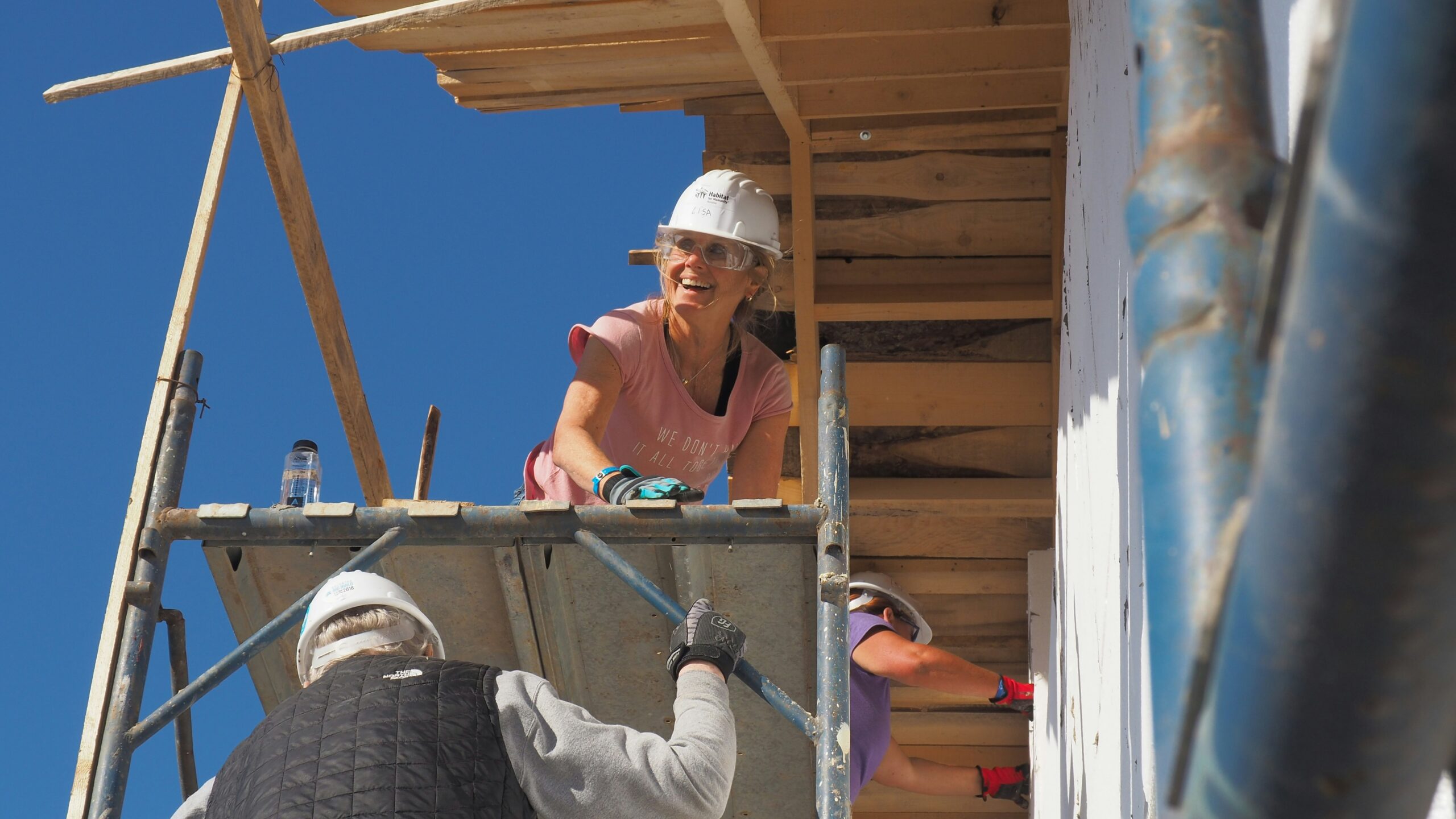 A woman in a hard hat kneels on a scaffold as she smiles down to someone climbing up the scaffold to her.