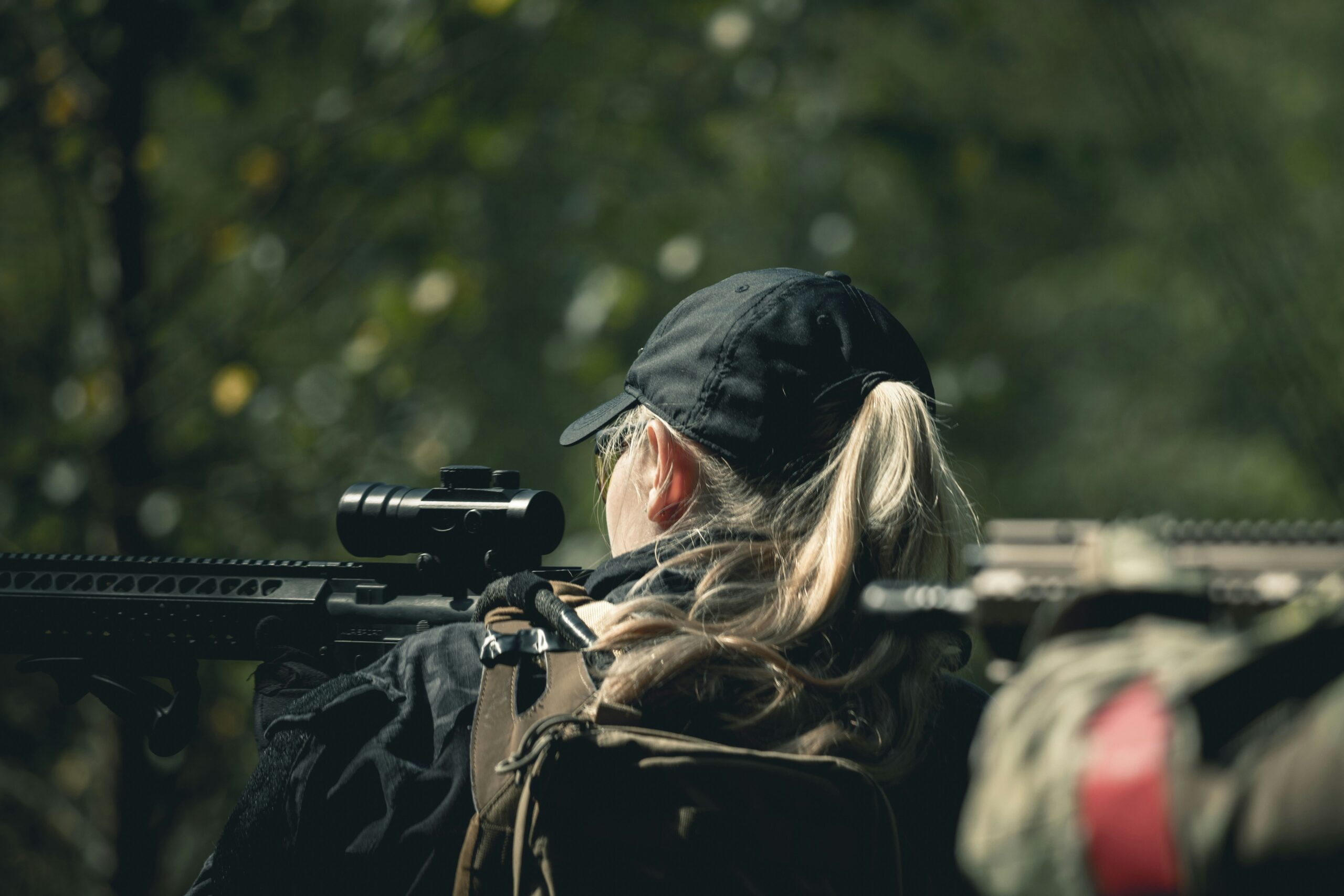 A woman with a backpack and a baseball cap stands with her rifle raised in the forrest.