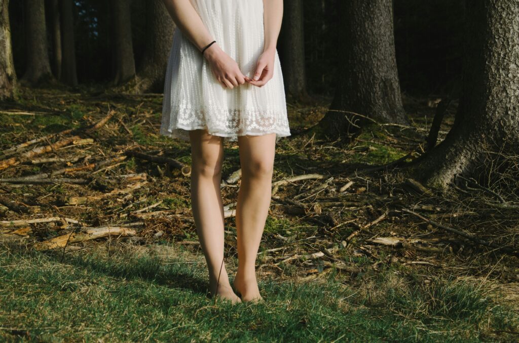 A woman in a white sundress stands in the forrest, her bare feet in the grass.