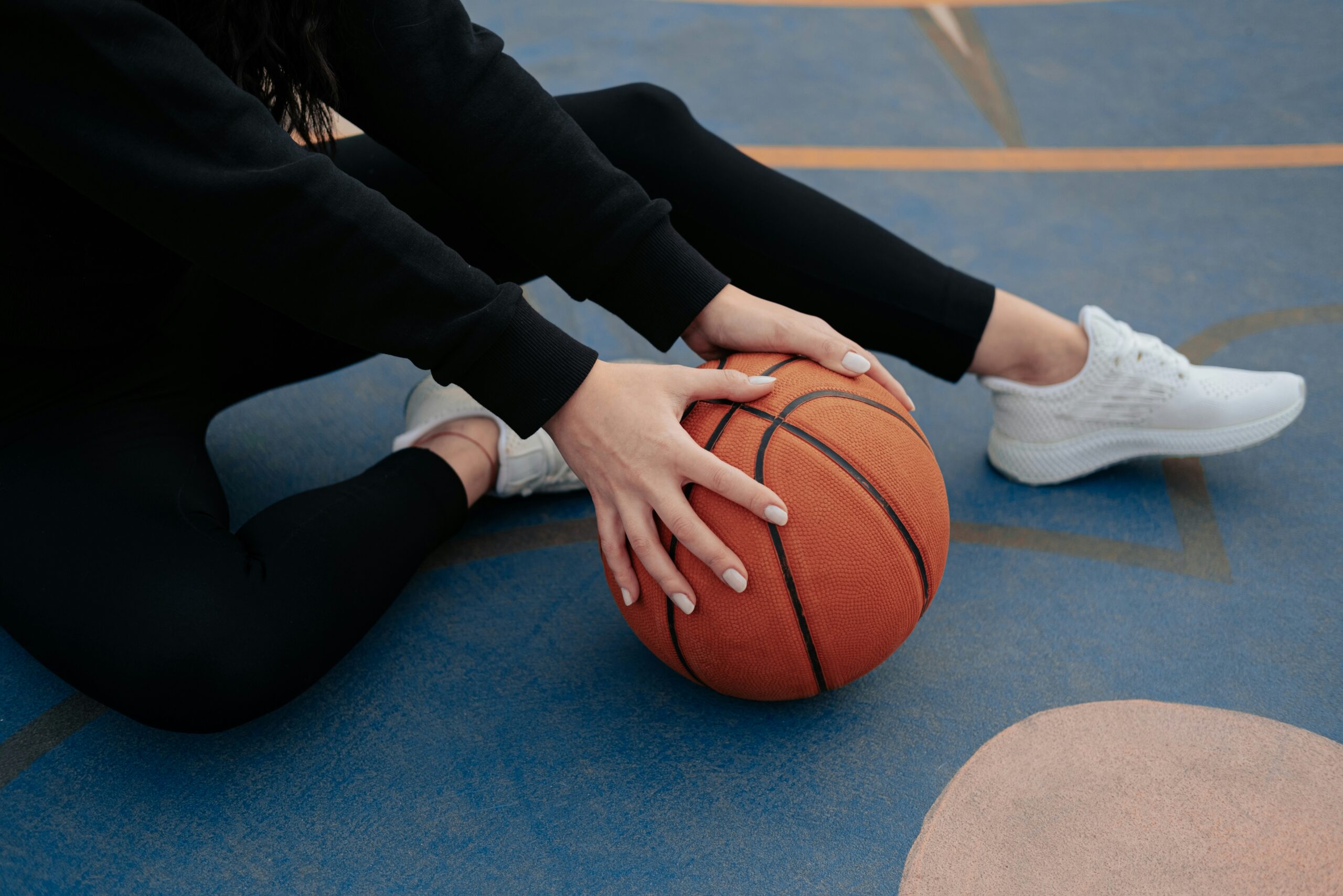 A woman sits on a basketball court. We see her legs and her hands and she holds a basketball on the ground.