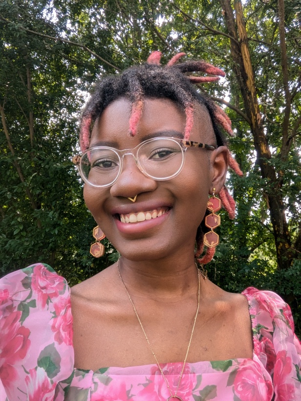 A Black person smiles as they look into the camera. Their hair is in dreads with pink tips at the ends.