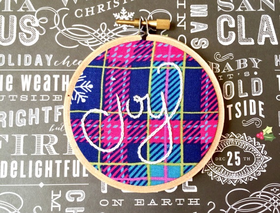 The word joy embroidered on a hot pink and blue plaid background, framed in a circular embroidery hoop